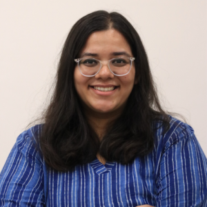 Smiling woman with glasses, long dark hair, wearing a blue striped shirt, standing against a plain background.