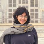 Smiling person with glasses and a scarf in front of an ornate building with detailed carvings.