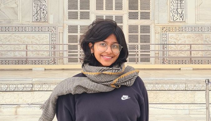 Smiling person with glasses and a scarf in front of an ornate building with detailed carvings.