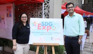 Two people standing next to an SDG Expo sign with red tents and event booths in the background.