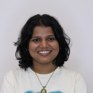 Smiling person with short wavy hair, wearing a white shirt and necklace, against a plain background.