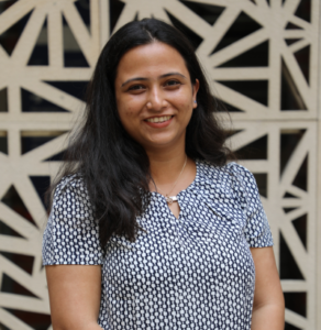 Smiling woman with long dark hair wearing a patterned blouse, standing in front of a geometric wall.