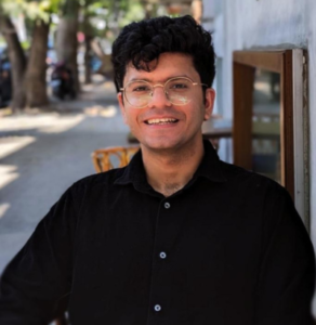Smiling person with glasses, black shirt, curly hair, standing outdoors near a tree-lined street.