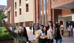 Group of people smiling and posing for a selfie outside a brick building with greenery nearby.