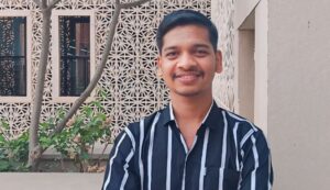 Smiling man in a black-and-white striped shirt standing outdoors near a patterned wall and plants.