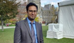 Man in a suit and tie standing outdoors on a grassy area with a historic building in the background.