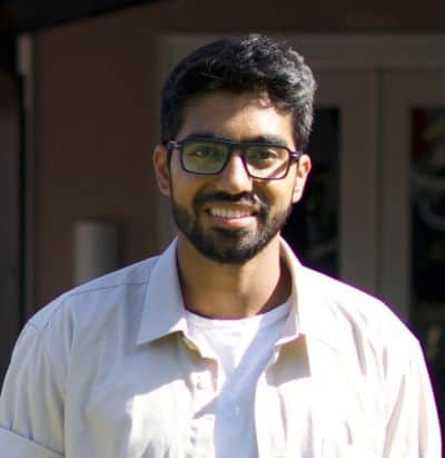 Smiling man with glasses, beard, and light-colored shirt standing outdoors in soft sunlight.