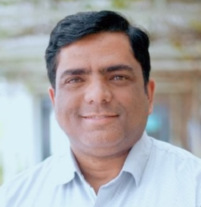 Smiling man with short black hair wearing a light blue shirt, blurred outdoor background.