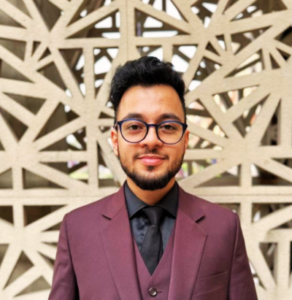 Man in a maroon suit and black shirt, wearing glasses, standing in front of a geometric patterned wall.