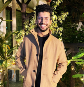 Smiling man in a beige coat with glasses, standing outdoors surrounded by plants and string lights.