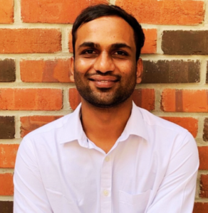 Smiling man in a white shirt standing in front of a brick wall.