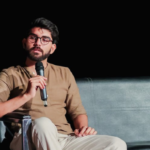 Man wearing glasses and a brown shirt, sitting on a black couch, holding a microphone.
