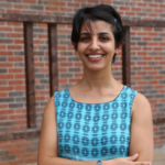 Smiling woman in a blue patterned dress, standing in front of a brick wall with wooden beams.