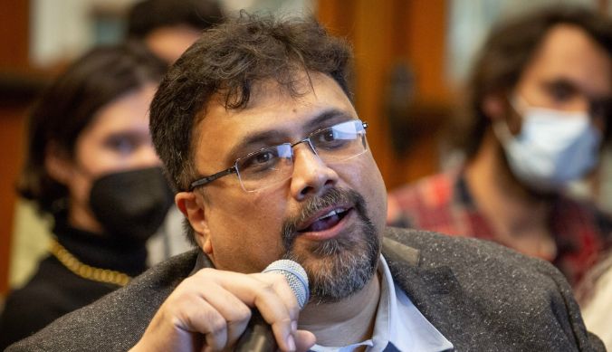 Man with glasses speaking into a microphone, surrounded by an audience, some wearing masks.