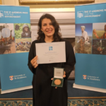 Woman smiling, holding a certificate and medal, standing between two St Andrews Prize banners.