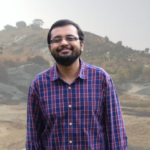 Smiling man in a red and blue checkered shirt standing outdoors with a rocky hill in the background.