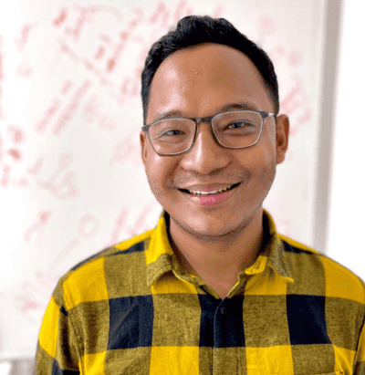 Smiling person wearing glasses and a yellow-black checkered shirt, whiteboard in the background.