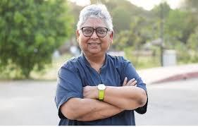 Smiling person with short gray hair, glasses, and a green watch, standing outdoors with arms crossed.
