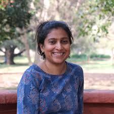 Smiling woman in a blue patterned top sitting outdoors with trees and greenery in the background.
