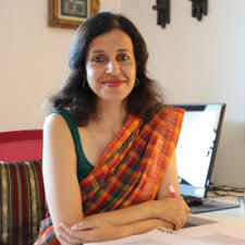 Woman in a colorful saree sitting at a desk with papers, smiling, and a computer in the background.