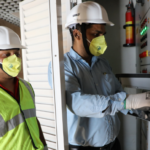 Two workers wearing helmets and masks inspect an electrical panel, one in a safety vest.