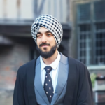 Man in formal attire with a checkered turban, standing outdoors near brick and timber buildings.