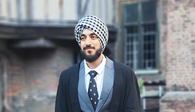Man in formal attire with a checkered turban, standing outdoors near brick and timber buildings.