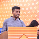 Man speaking at a podium with a decorative orange and yellow patterned backdrop.
