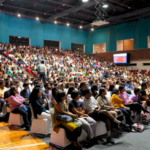 Large audience seated in an auditorium with teal walls, bright lights, and two projection screens.
