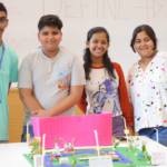 Four students standing behind a colorful model project on a table, with a whiteboard in the background.