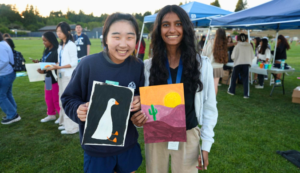 Two people smiling and holding paintings, one of a goose and the other of a desert scene with a cactus.