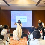 Speaker addressing an audience in a conference room with an Ashoka University presentation on screen.