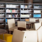 Library with bookshelves, wooden tables, and cream-colored chairs in a well-lit space.