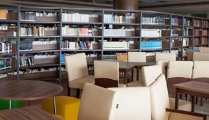 Library with bookshelves, wooden tables, and cream-colored chairs in a well-lit space.