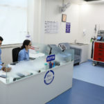 Healthcare reception desk with two staff members, a hospital bed, and medical equipment in the background.