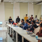 Person speaking to an attentive audience in a classroom with desks, laptops, and seated participants.