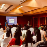 Audience seated in a conference room, speakers presenting near a screen displaying "Ashoka".