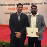 Two men stand on a red-carpeted stage holding a certificate at an award ceremony with flags behind them.