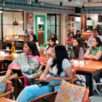 People seated in a cafe-style room, attentively listening, with tables and a counter in the background.