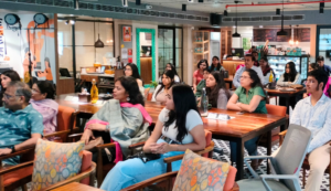 People seated in a cafe-style room, attentively listening, with tables and a counter in the background.