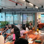 Speaker addressing a seated audience in a modern coworking space with plants and wall art.
