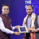 Two men exchanging a plaque on stage, one wearing a vest and the other wearing a patterned scarf.
