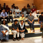 Group of people seated on tiered wooden steps in a well-lit room with geometric wall patterns.