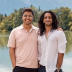 Two men standing by a lake with trees, mountains, and an ornate fence in the background.