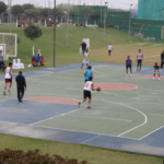 Outdoor basketball game in progress, players in white and blue jerseys, spectators on the sidelines.