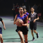 A basketball player in a blue jersey drives toward the hoop, pursued by opponents on an outdoor court.