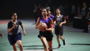 A basketball player in a blue jersey drives toward the hoop, pursued by opponents on an outdoor court.