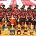 A sports team in red and black jerseys posing with multiple gold trophies and awards in a stadium.