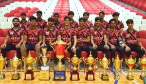 A sports team in red and black jerseys posing with multiple gold trophies and awards in a stadium.