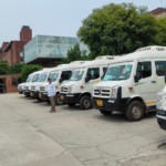 Row of white vans parked outside a modern brick and glass building with trees in the background.